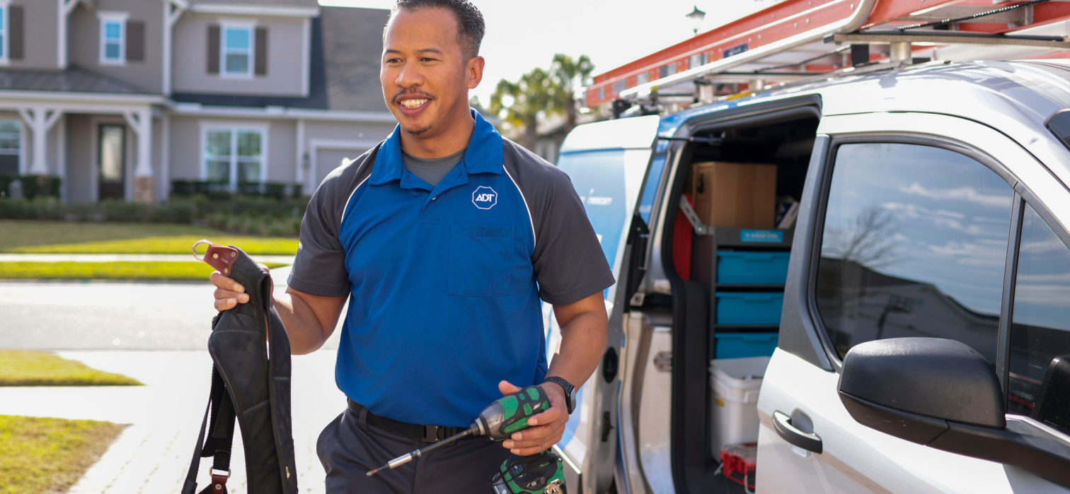A male ADT Field Operations employee holds a drill and tool belt as he stands next to his van in a driveway.
