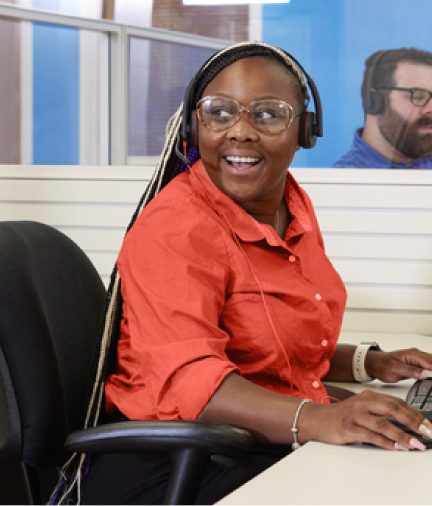A young woman wearing a microphone headset and working at her computer, smiles as she turns to talk to someone.